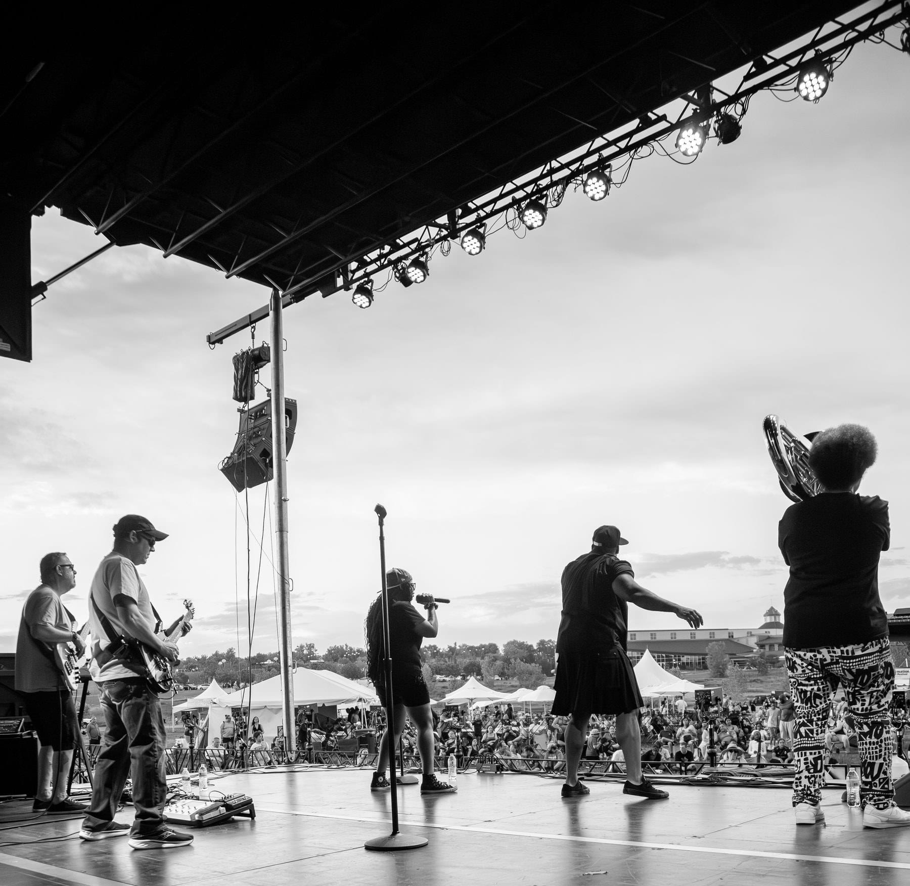 The band on the festival stage at Juneteenth in Thornton, seen from behind the drums with the crowd beyond, in black and white.
