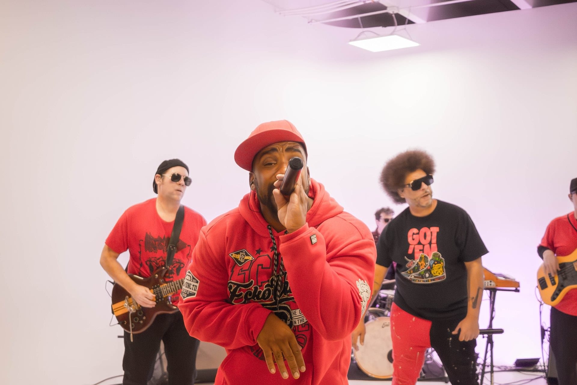 A singer in a red hoodie on the microphone with the band behind.