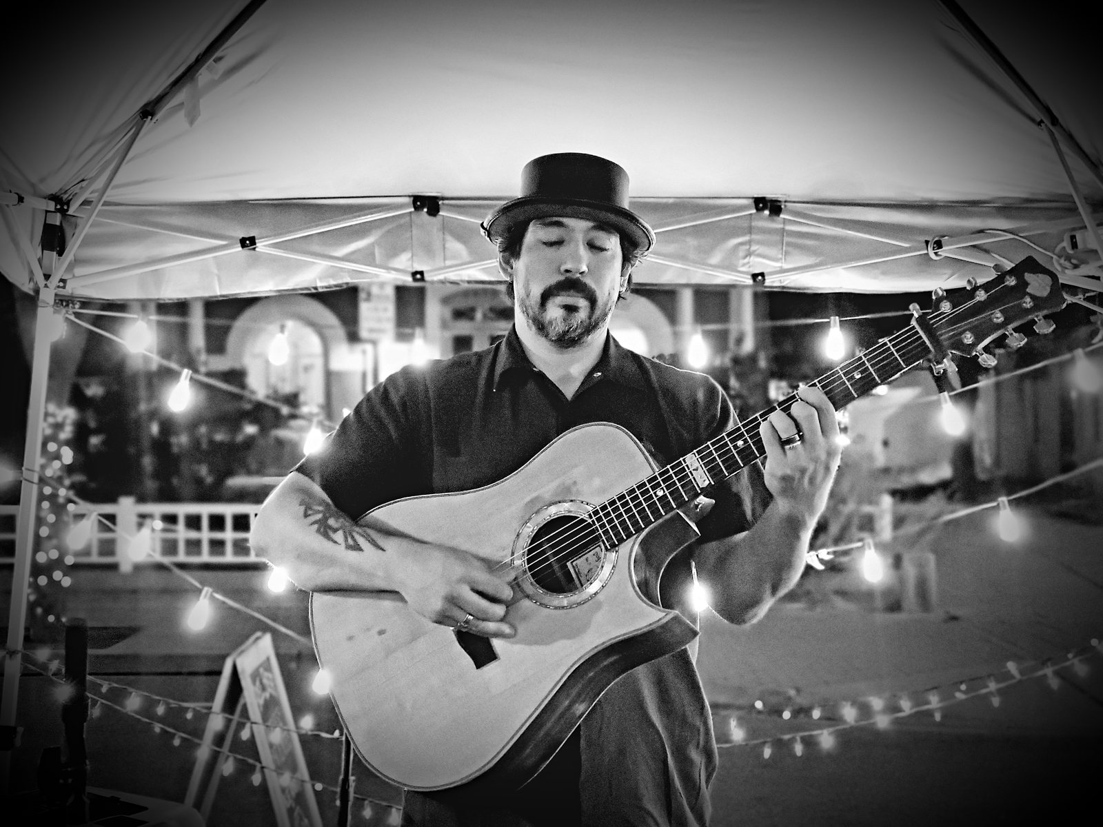 Tony Medina playing an acoustic guitar under string lights, black and white.