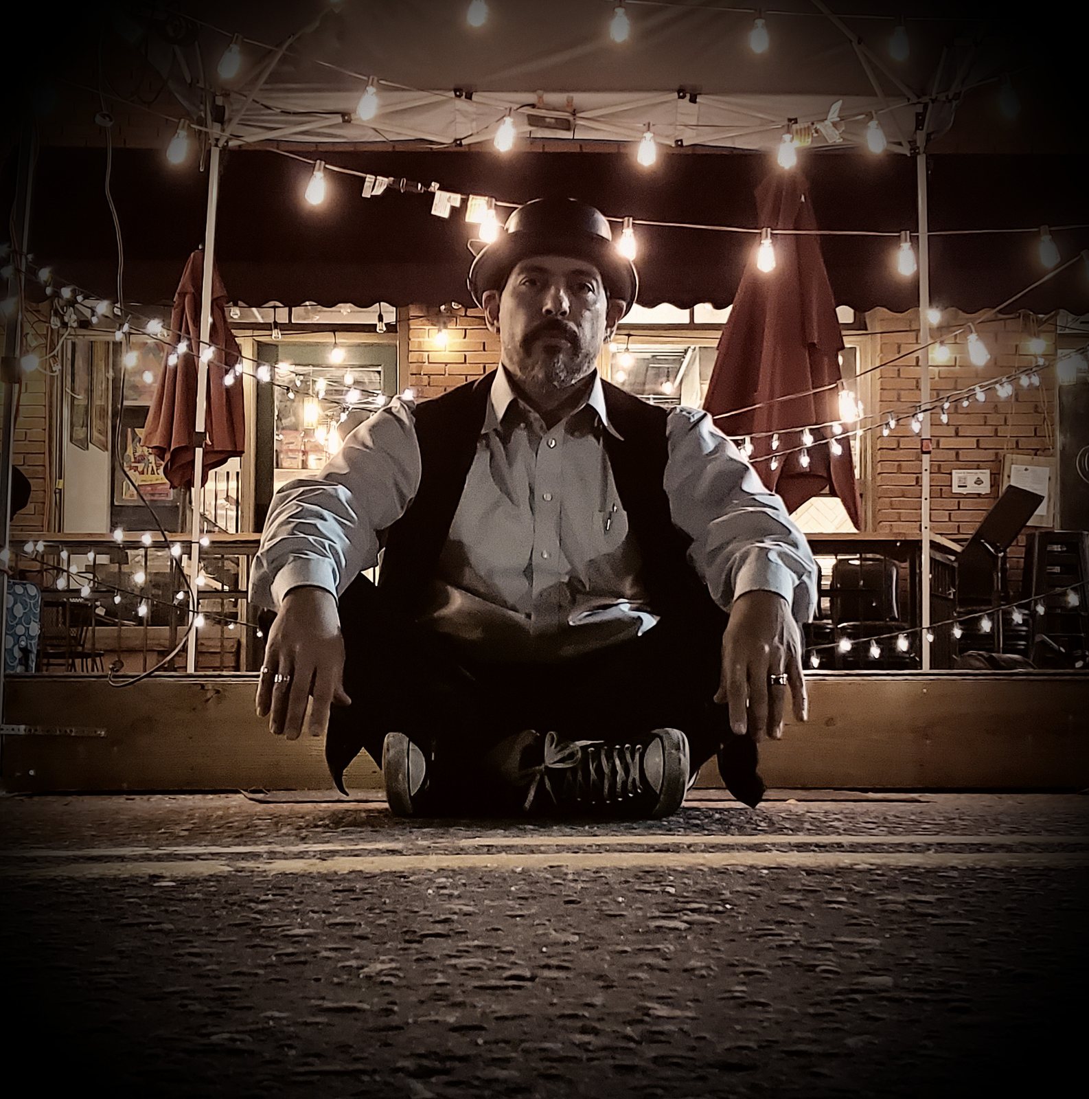 Tony Medina seated on cobbles in a hat and waistcoat, a lit patio behind him.