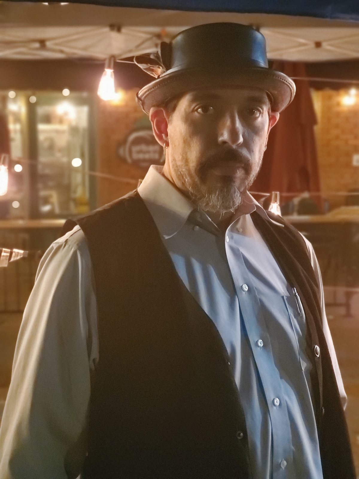 Tony Medina in a hat and waistcoat outside a bar at night.