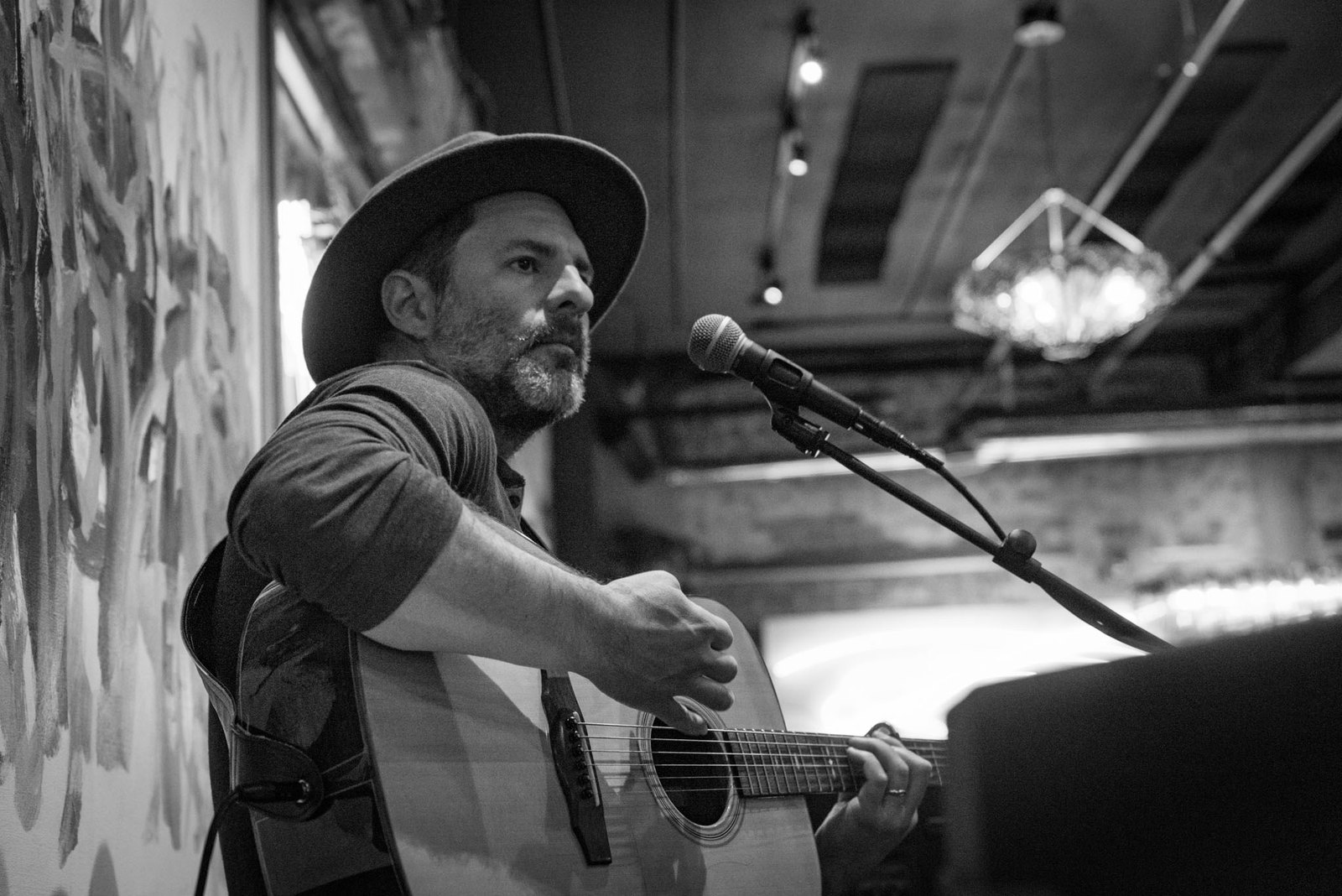 René Moffatt playing an acoustic guitar in front of a large abstract painting, in black and white.