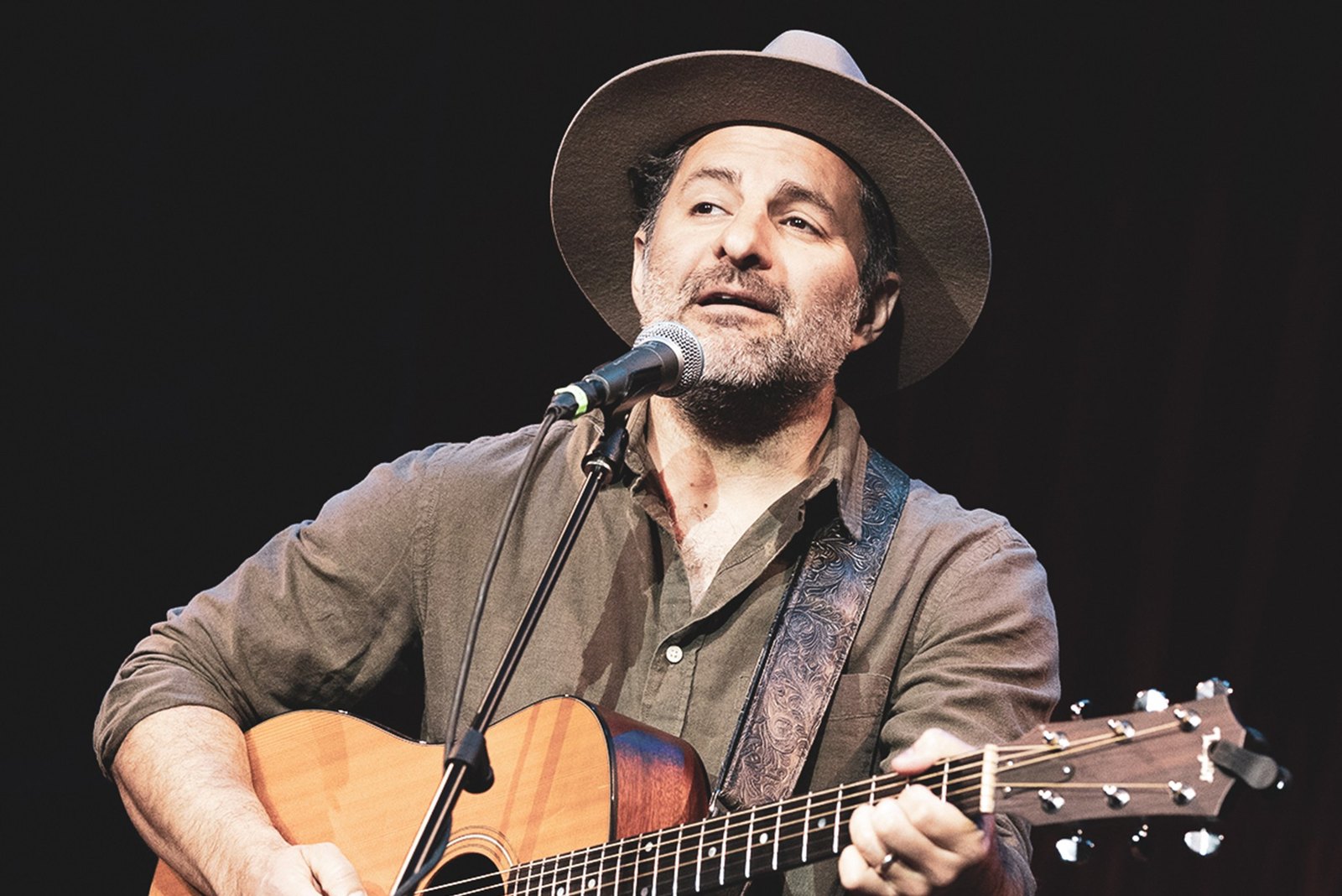 René Moffatt singing into a microphone on a festival stage, guitar strap over his shoulder.
