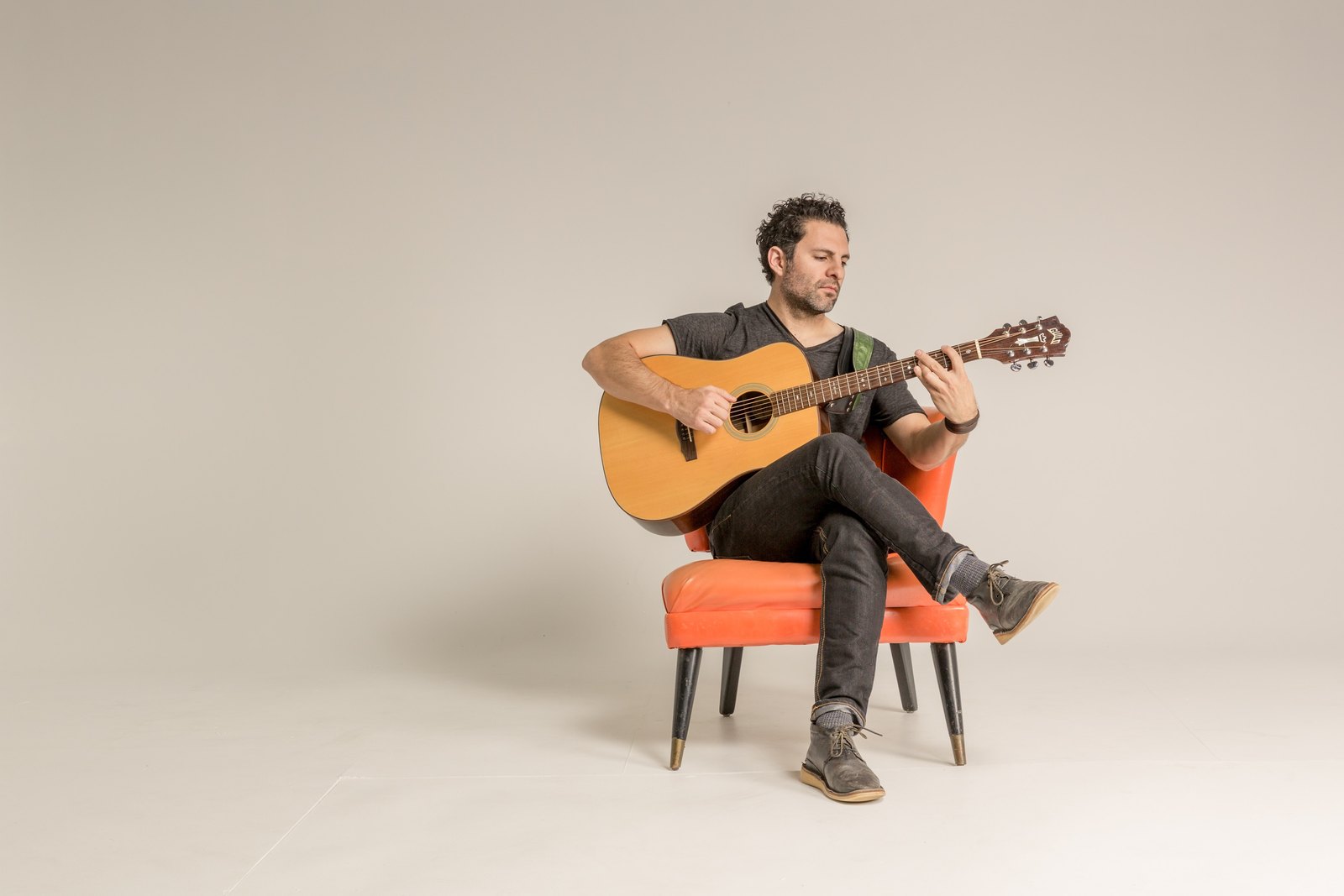 René Moffatt seated in an orange armchair with an acoustic guitar against a plain backdrop.