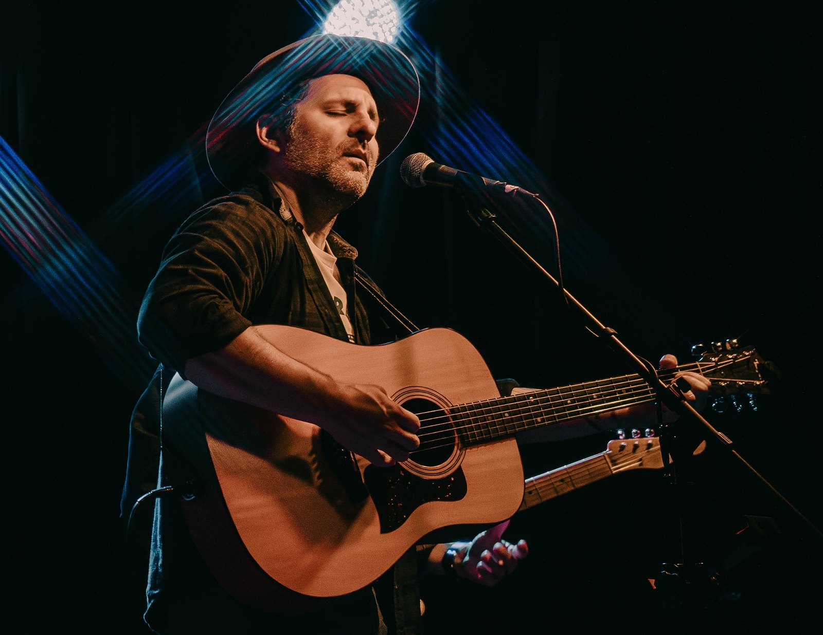 René Moffatt singing at a microphone with an acoustic guitar, in a hat under warm stage light.