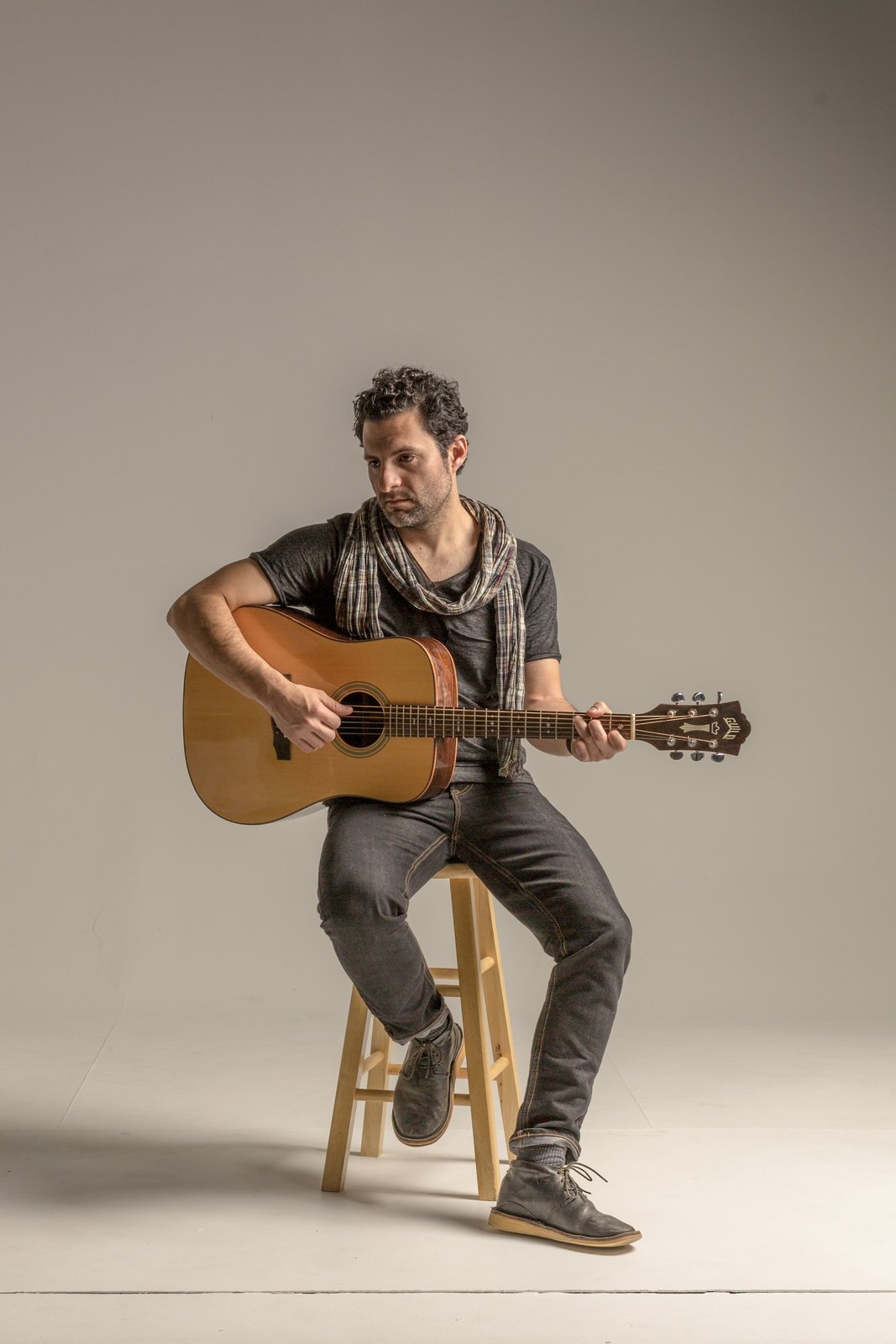 René Moffatt on a stool playing an acoustic guitar, in a scarf, against a white backdrop.