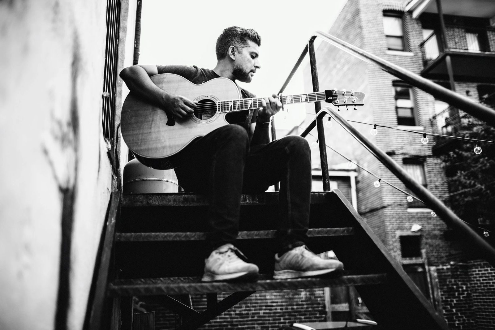 Tejas Singh playing an acoustic guitar on a fire escape, in black and white.
